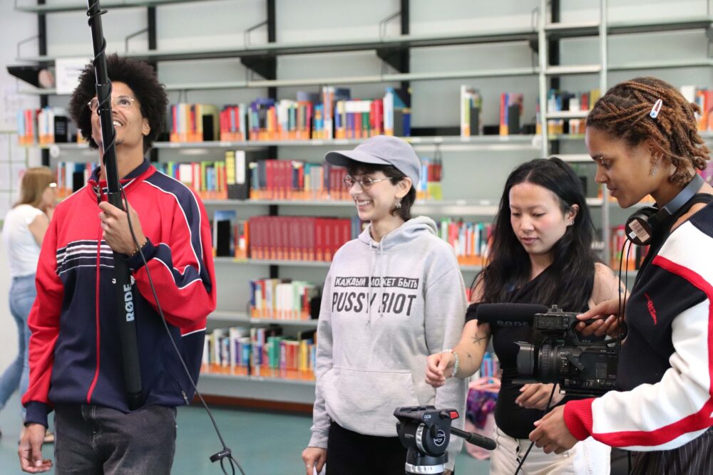 4 Personen stehen nebeneinander vor einem großen Bücherregal. Links hält eine Person ein Mikrofon, die Personen rechts stellen Kamera und Stativ ein. Alle schauen fröhlich.
