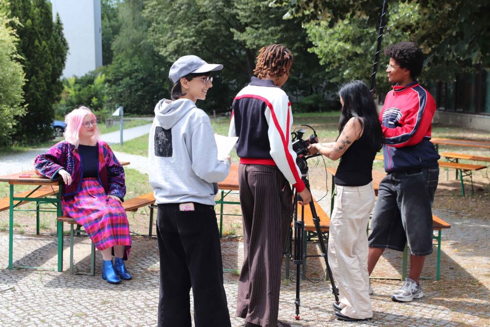 Links im Bild sitzt eine Person draußen auf einer Bierbank. 3 Personen stehen um eine Videokamera und nehmen diese Person auf. Eine Person schaut lächelnd nach rechts und hat Zettel in der Hand.