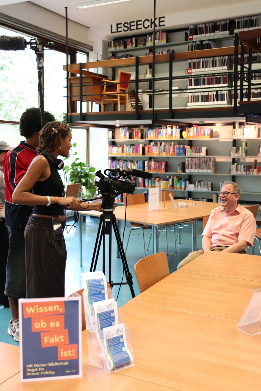 Eine Person sitzt in einer Bibliothek auf einem Stuhl. 2 Personen sthene hinter einer Videokamera und nehmen diese Person auf.