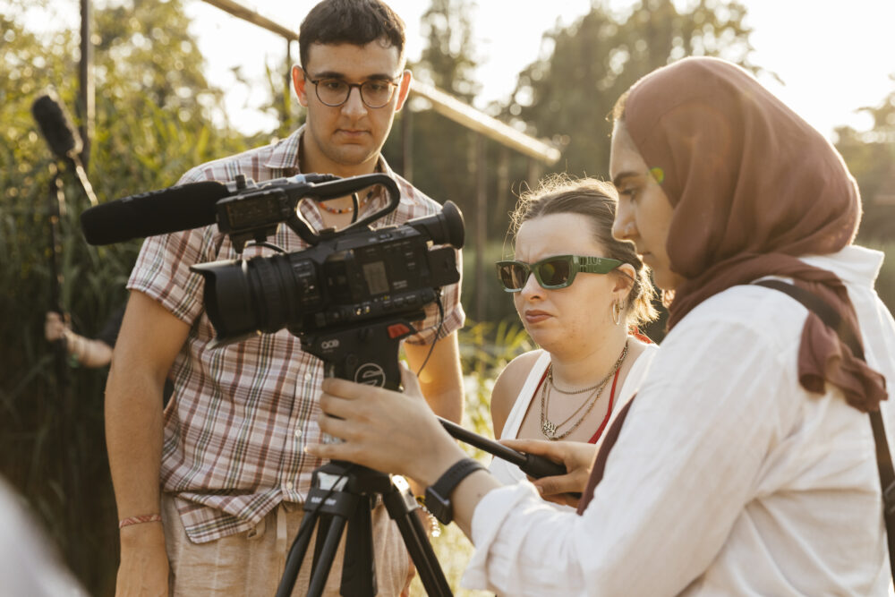 3 Personen schauen auf eine Videokamera mit Mikrofon. Im Hintergrund sind Bäume. Die Sonne wirft ein warmes Licht.