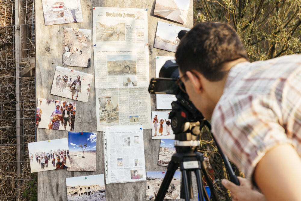 Eine Person macht Nahaufnahmen mit einer Videokamera von einer Wand mit Fotos und Zeitungsartikeln.