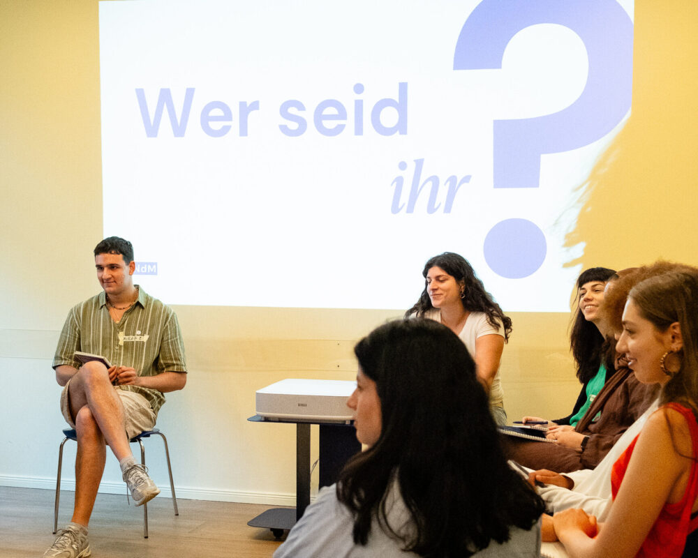 Die Teilnehmenden des Projekts sitzen im Kreis, der Beamer strahlt den Satz "Wer seid ihr?" im Hintergrund an die Wand.