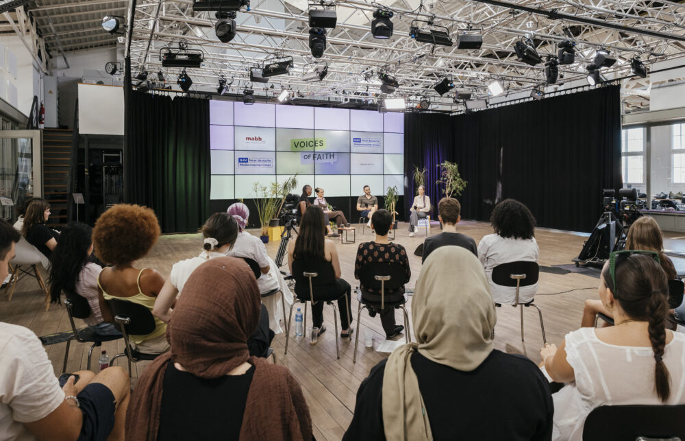 Ein Publikum schaut ein Panel im Studio an. Im Panel sitzen 4 Personen vor einem Hintergrund mit Logos.