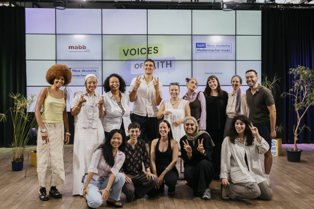 Gruppenfoto im Studio von ALEX. In der hinteren Reihe stehen 9 Personen, vor ihnen hocken 5 Personen. Im Hintergrund ist das Set für die Sendung "Voices of Faith". Viele machen lustige Handzeichen.