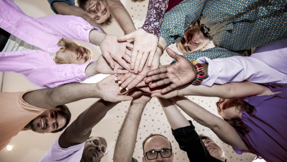 Das Foto zeigt mehrere Personen, die im Kreis stehen und ihre Hände als Zeichen von Gemeinschaft übereinander legen.