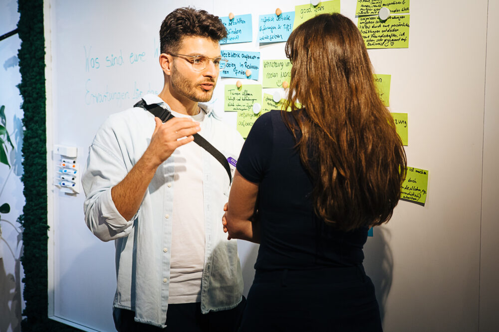 Das Bild zeigt zwei Personen, die auf einer Veranstaltung des Mentoring@NRW vor einem Whiteboard stehen. Eine Person steht mit dem Rücken zur Kamera. Die Personen unterhalten sich. An dem Whiteboard sind Notizzettel magnetisch angepinnt.