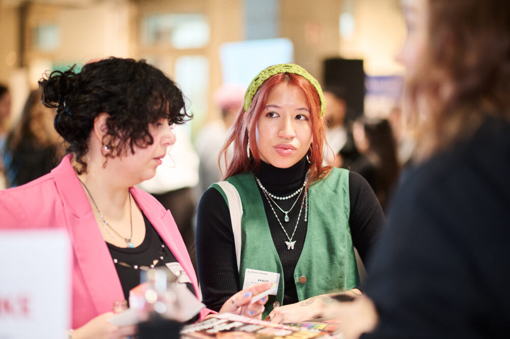 Auf dem Bild sind zwei Personen mit Namensschildern auf ihrem Oberteil zu sehen. Sie stehen vor einem Stand mit Stickern und Karten und unterhalten sich miteinander.