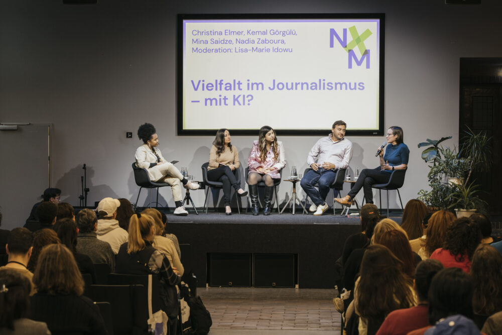 Das Bild zeigt ein Panel mit fünf Personen bei der NxM 2024. Auf einer Leinwand im Hintergrund in der Titel der Paneldiskussion zu sehen: "Vielfalt im Journalismus - mit KI?"