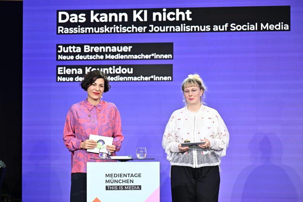 Zwei Mitarbeiterinnen der Neuen deutschen Medienmacher*innen stehen als Referentinnen vor einer Leinwand mit dem Vortragstitel „Das kann KI nicht – Rassismuskritischer Journalismus auf Social Media“ bei den Medientagen München.