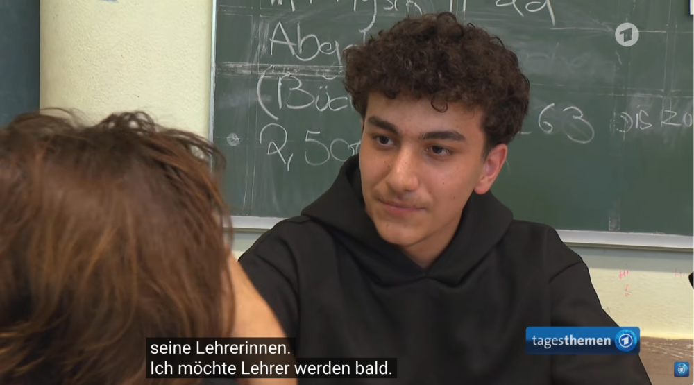 Journalistin Isabel Schayani sitzt mit dem Schüler Bilal in einem Klassenzimmer. Er erzählt, dass er bald Lehrer werden möchte. Bilal trägt einen schwarzen Hoodie.