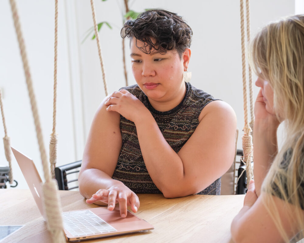 Mentee und Workshopleiterin sitzen an einem Holztisch, eine zeigt auf den Bildschirm eines Laptops, die andere hört aufmerksam zu