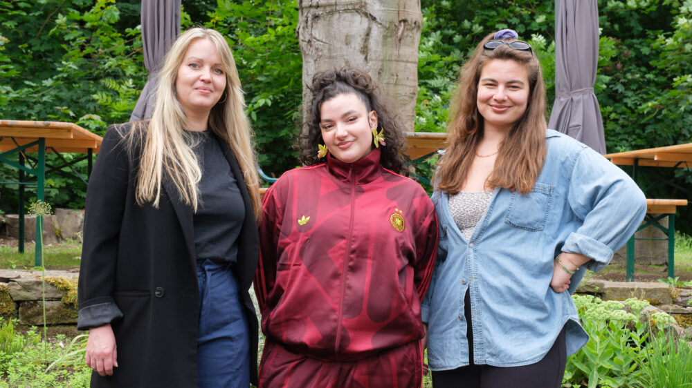 Drei Frauen (Workshopleiterinnen) stehen vor einem Baum im Freien, die linke trägt schwarzen Blazer und blaue Jeans, die mittlere einen roten freshen Trainingsanzug, die rechte eine blaue Bluse und hat die Hand in der Hüfte.