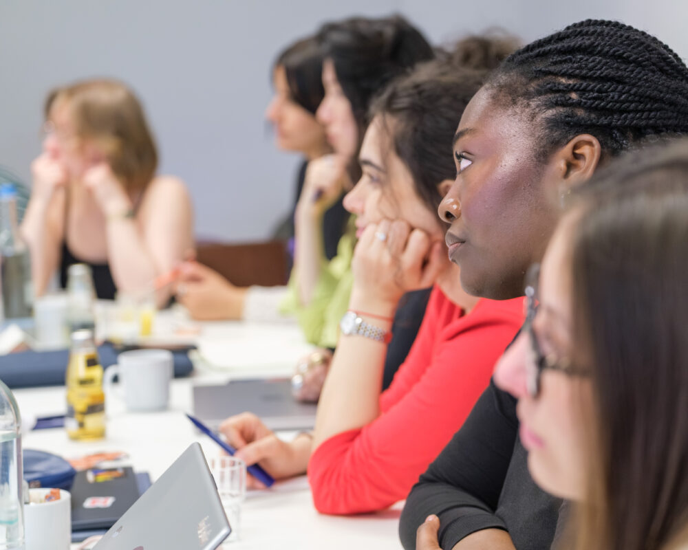 Mehrere junge Frauen (Mentees) sitzen in einem Workshop des NRW Mentoring Camps an einem Tisch und hören aufmerksam zu
