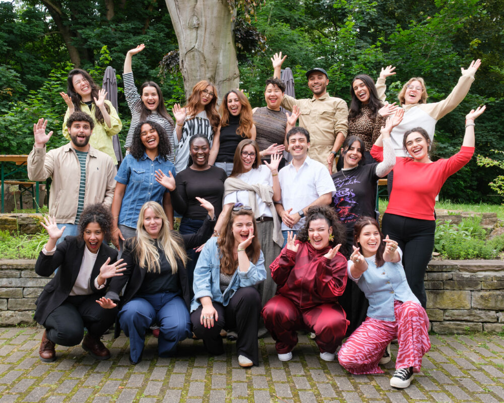 Gruppenbild mit fröhlichen Teilnehmer*innen und Workshopleiterinnen des NRW Mentoring Camps vor einem Baum und einer Steinmauer, die Hände winkend und lachend in die Kamera schauen
