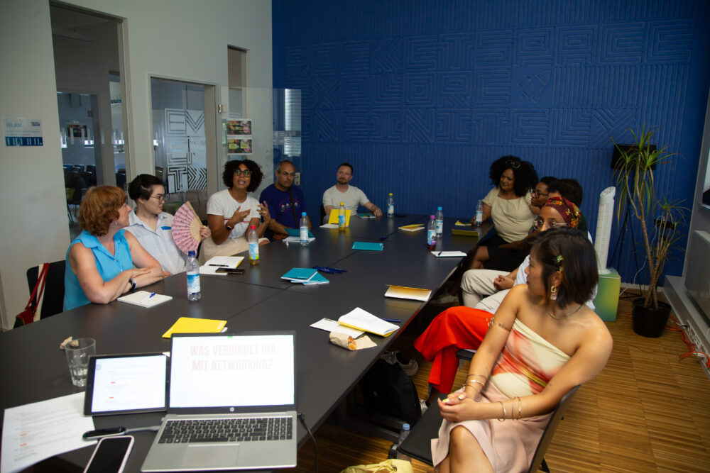 Eine Gruppe von Menschen sitzen an einem langen Tisch vor einer blauen Wand. Viele von ihnen sind BIPoC. Es sind Workshopteilnehmende des Events Take your Space.