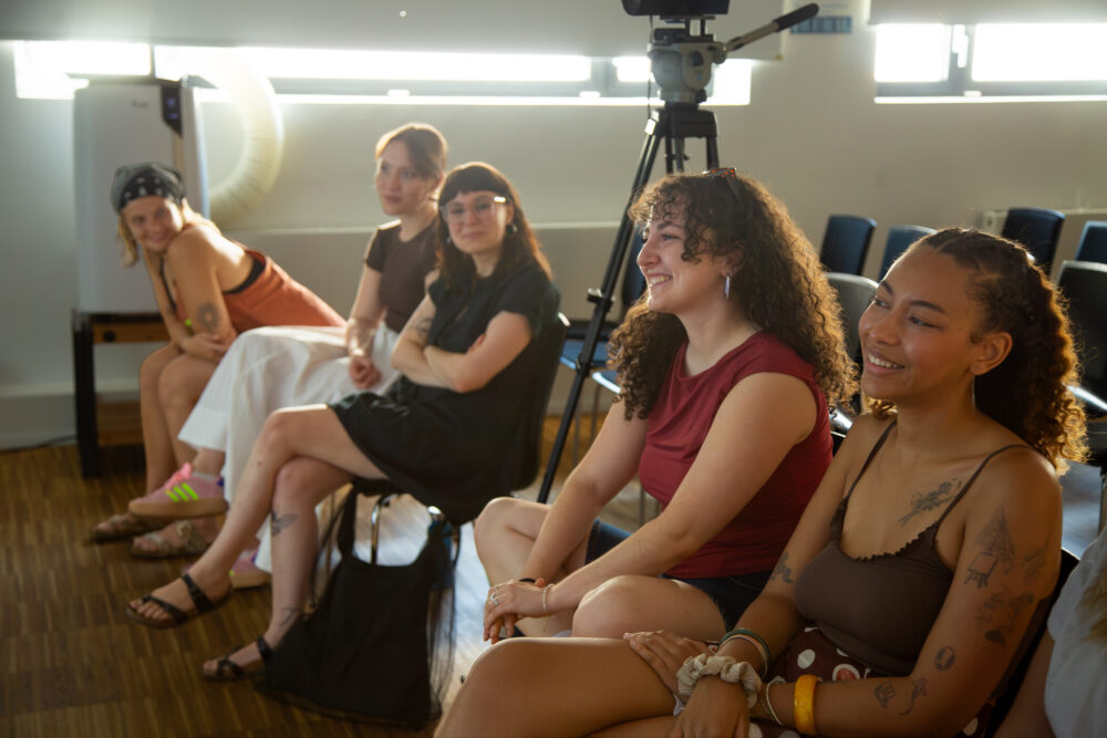 Mehrere junge Menschen sitzen gut gelaunt nebeneinander in einem Konferenzraum. Sie tragen sommerliche Kleidung. Im Hintergrund ist eine mobile Klimaanlage zu sehen.