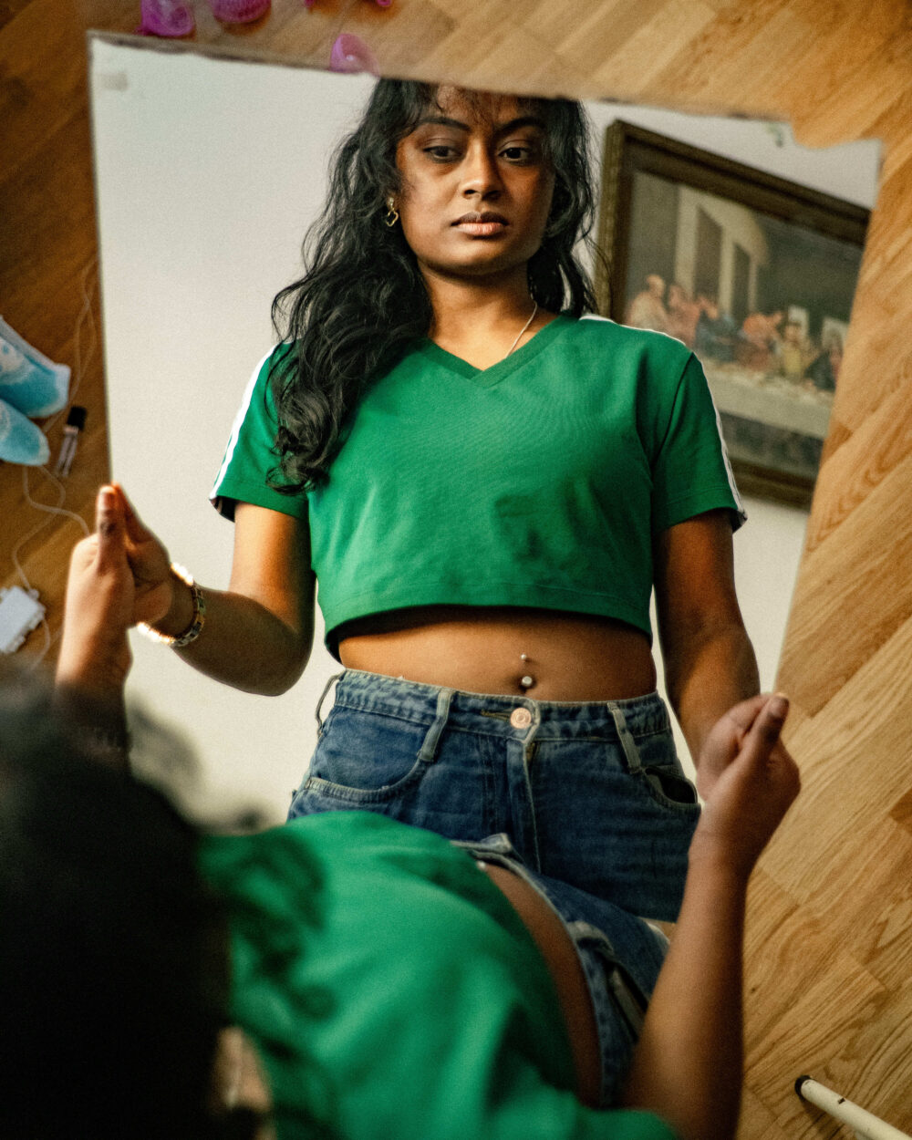 Das Bild zeigt eine junge Frau, die in ihr Spiegelreflexion schaut. Sie trägt ein grünes Crop-Top mit weißen Streifen an den Ärmeln, eine Jeans und ein Bauchnabelpiercing, dazu eine Halskette und ein Armband. Ihre Hände sind leicht ausgestreckt, ihr Blick ist ruhig und direkt in den Spiegel gerichtet. Im Hintergrund des Spiegelbilds ist ein angeschnittener Druck von Leonardo da Vincis „Das letzte Abendmahl" an der Wand zu erkennen, was auf ihren Wohnort in einer reformierten Kirche hinweist. Die Aufnahme ist leicht schräg und durch den Spiegelrahmen gerahmt, was der Komposition eine besondere Tiefe verleiht.