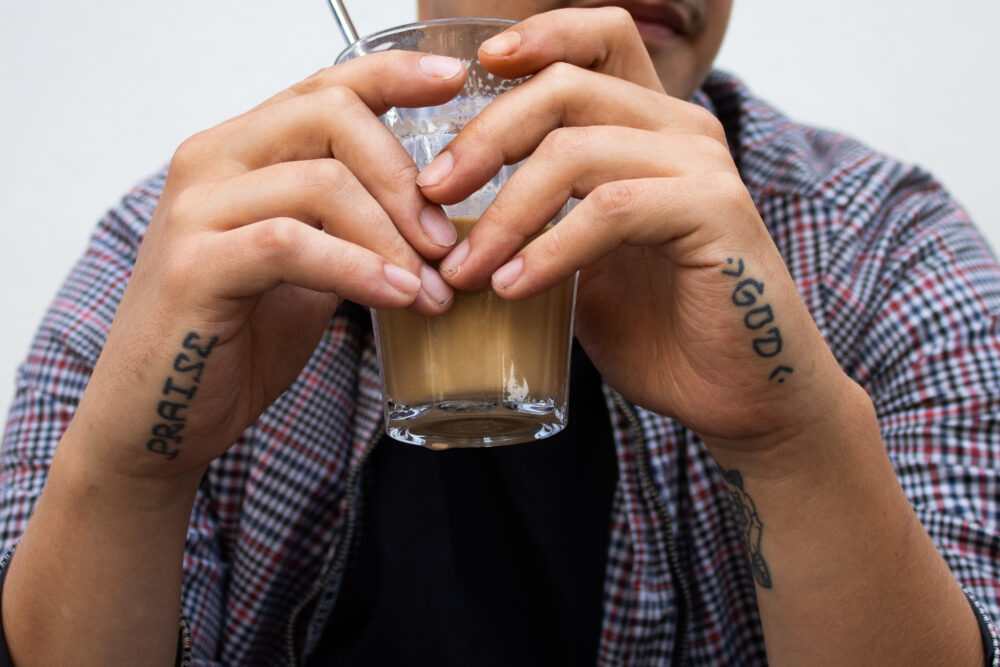 Nahaufnahme von zwei Händen, sie halten ein Glas Kaffee. An den Handseiten sind jeweils Tattoos, auf denen "Praise" und "God" steht.