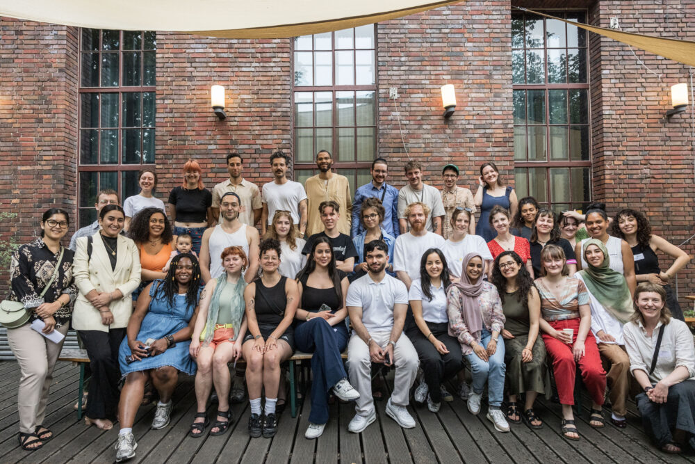 Eine größere, vielfältige Gruppe junger Menschen sitzt und steht dicht beieinander vor einer Backsteinfassade im Freien und blickt in die Kamera, als gemeinsames Gruppenfoto bei der Auftaktveranstaltung vom Projekt Beyond the Frame.