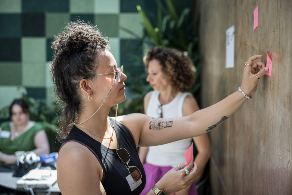 Bei einem Workshop klebt eine Person mit Brille und Zopf mit dem linken Arm ein Post-it an die Wand; im Hintergrund stehen und sitzen zwei weitere Personen an einem Tisch.