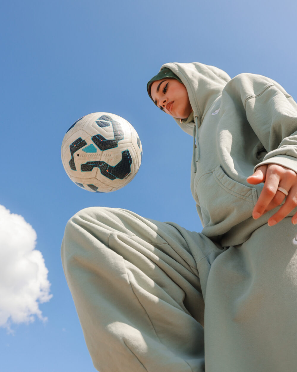 Das Bild zeigt eine junge Frau mit Kopftuch, die beim Fußballspielen festgehalten wird. Die Aufnahme erfolgt aus einer Untersicht, wodurch ihr Bein und der Ball im Vordergrund betont werden. Sie jongliert den Ball mit dem Oberschenkel, während ihr Blick nach unten auf den Ball gerichtet ist. Die Frau trägt einen hellgrünen Trainingsanzug, der sich deutlich vom Hintergrund abhebt. Hinter ihr ist ein klarer blauer Himmel zu sehen, in dem sich eine einzelne Wolke befindet.