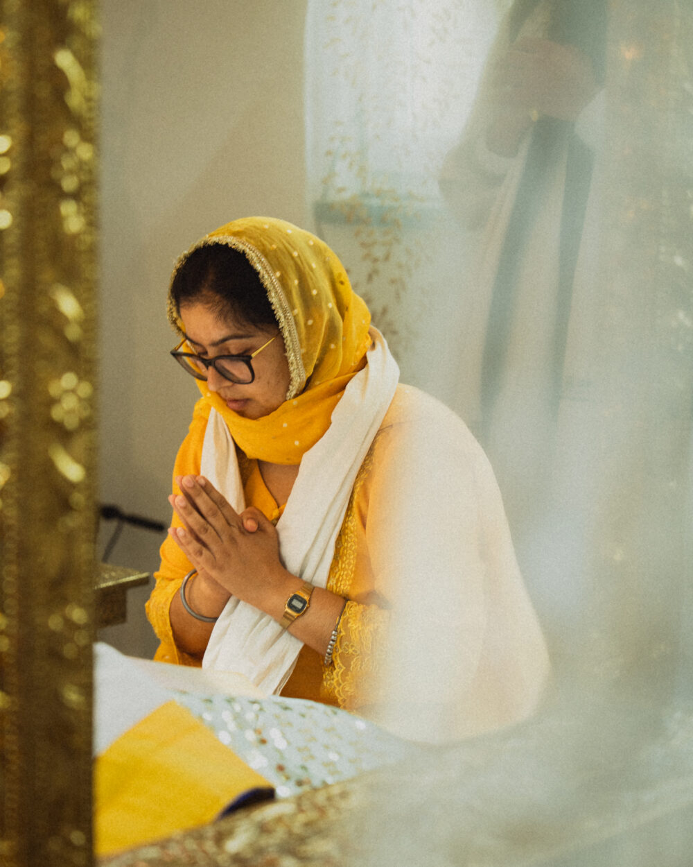 Das Bild zeigt eine Frau der Sikh-Gemeinde beim Gebet. Sie hat die Hände vor der Brust gefaltet und die Augen geschlossen. Sie trägt einen gelben Dupatta mit goldener Bordüre sowie einen weißen Schal, eine Brille und eine goldene Armbanduhr. Die Aufnahme erfolgt leicht von oben und seitlich, wobei ein verspiegeltes Element im Vordergrund die Komposition rahmt. Im Hintergrund sind goldene Verzierungen zu erkennen, die auf einen religiösen Raum hinweisen.