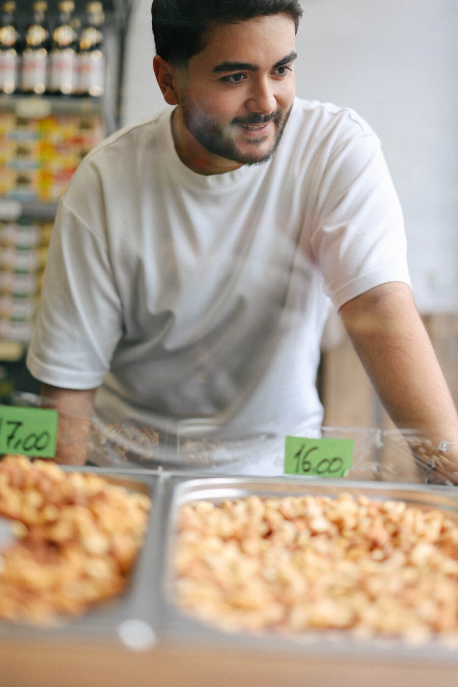 Das Bild zeigt einen jungen Mann hinter einer Verkaufstheke in einem Supermarkt. Er trägt ein weißes T-Shirt, lächelt und blickt leicht zur Seite. Im Vordergrund stehen zwei flache Metallschalen mit Nüssen, die mit grünen Preisschildern ausgezeichnet sind – eines zeigt 7,00, das andere 16,00. Im Hintergrund sind Regale mit Waren zu erkennen, die unscharf bleiben.
