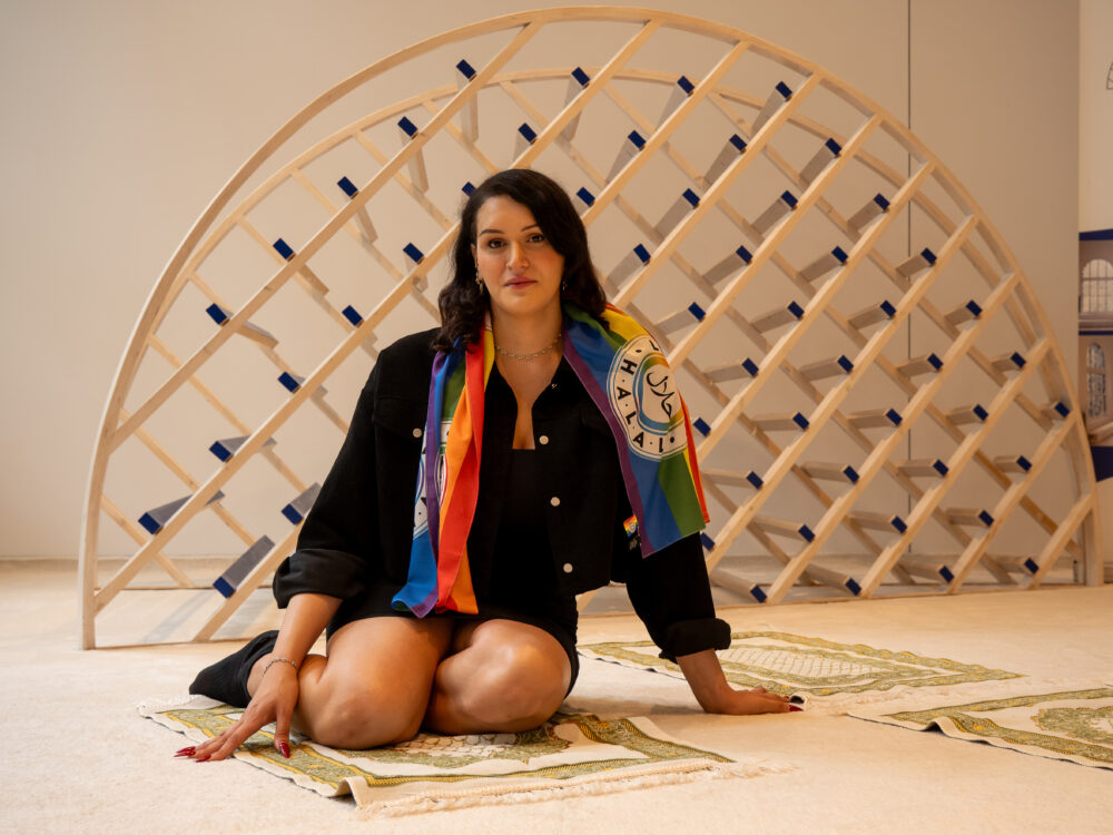 Das Bild zeigt eine Trans*-Frau, die auf einem Gebetsteppich sitzt und direkt in die Kamera blickt. Sie trägt eine schwarze Jeansjacke und einen Regenbogen-Schal mit dem Schriftzug „Halal" in arabischer und lateinischer Schrift. Hinter ihr ist eine halbkreisförmige Holzskulptur zu sehen – eine Qibla-Skulptur der Ibn Rushd-Goethe Moschee. Der Hintergrund ist hell und der Raum wirkt offen und licht.
