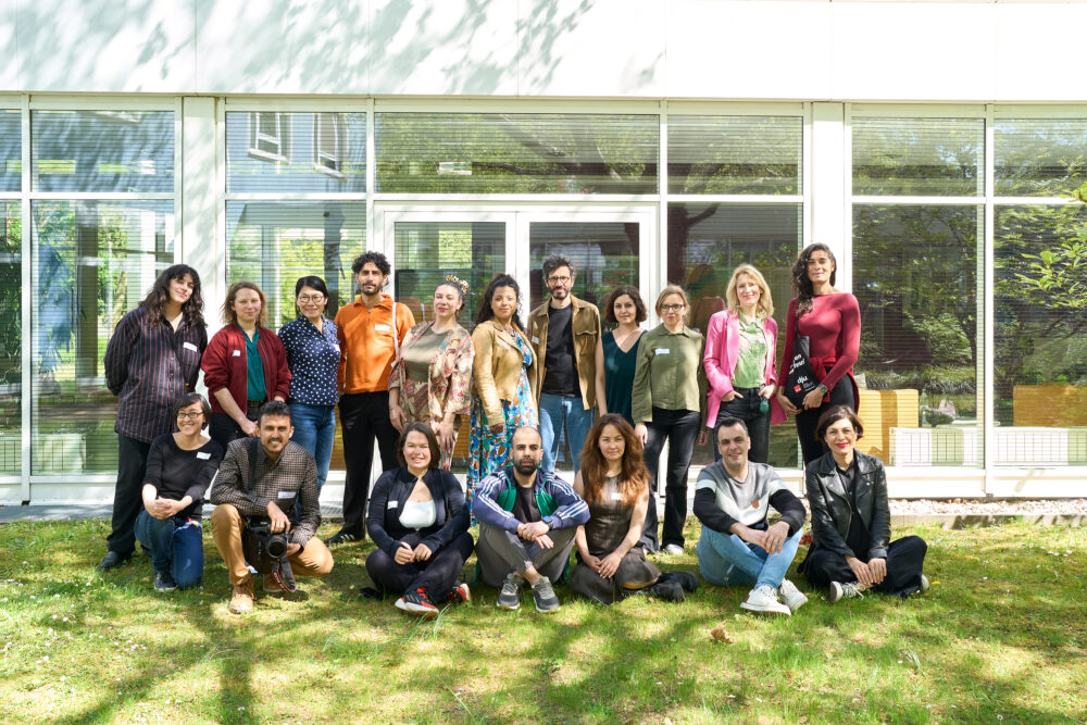 Eine Gruppe Erwachsener steht und sitzt auf einer Wiese vor einem Gebäude mit großen Glasfronten und blickt in die Kamera, als gemeinsames Gruppenfoto bei einer Veranstaltung.