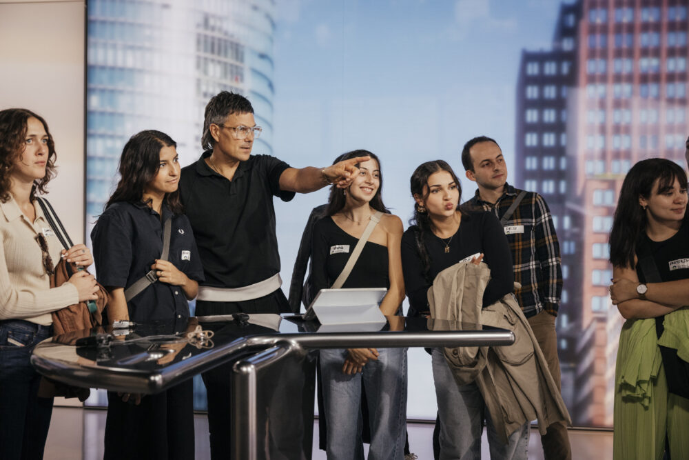 Sieben Veranstaltungsteilnehmer*innen stehen an einem Tisch. Eine Person zeigt auf etwas im Raum, das die anderen ansehen.