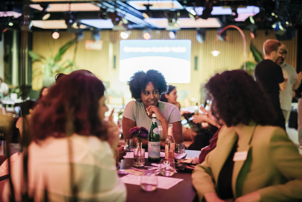 Das Foto zeigt drei Personen, die bei einer Veranstaltung um einen Tisch sitzen und sich unterhalten.