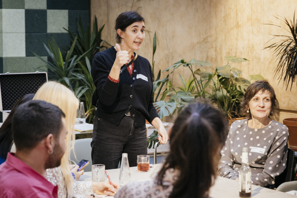 Das Bild zeigt eine Workshopsituation. Die Teilnehmer*innen sitzen um einen Tisch. Eine Person steht und erklärt etwas.