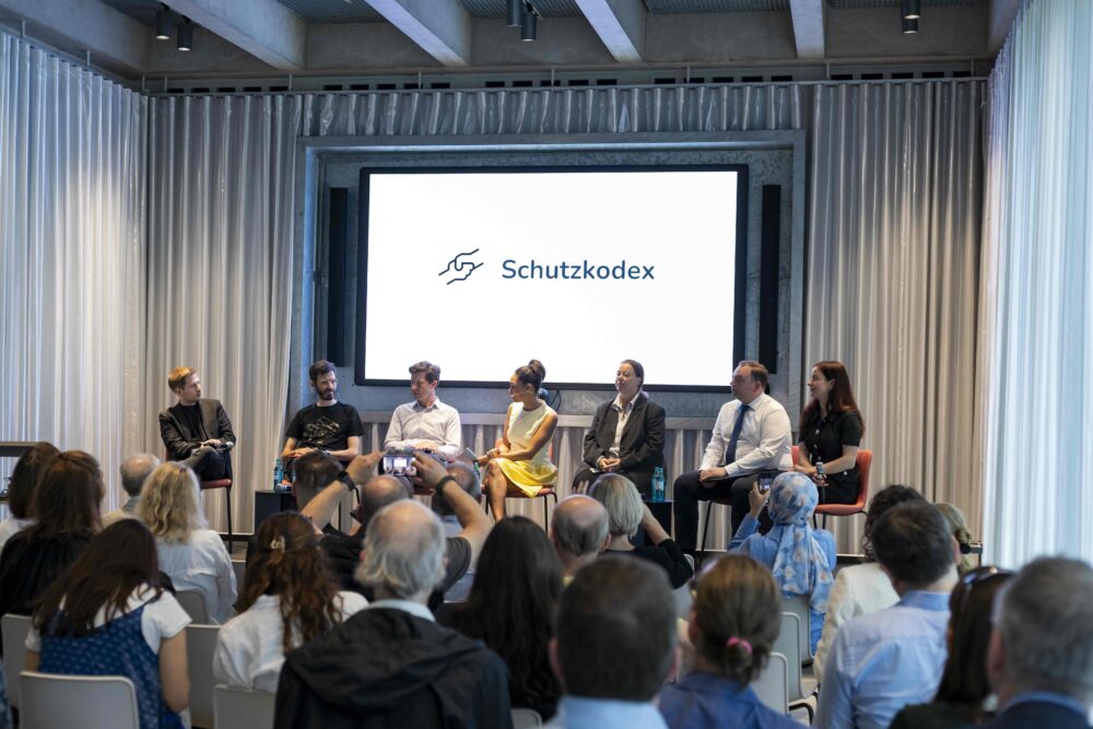 Das Bild zeigt eine Veranstaltung vom Schutzkodex. Sieben Personen sitzen auf einem Panel. Das Publikum sitzt mit dem Rücken zur Kamera. Auf der Leinwand hinter den Panelteilnehmer*innen steht „Schutzkodex“.
