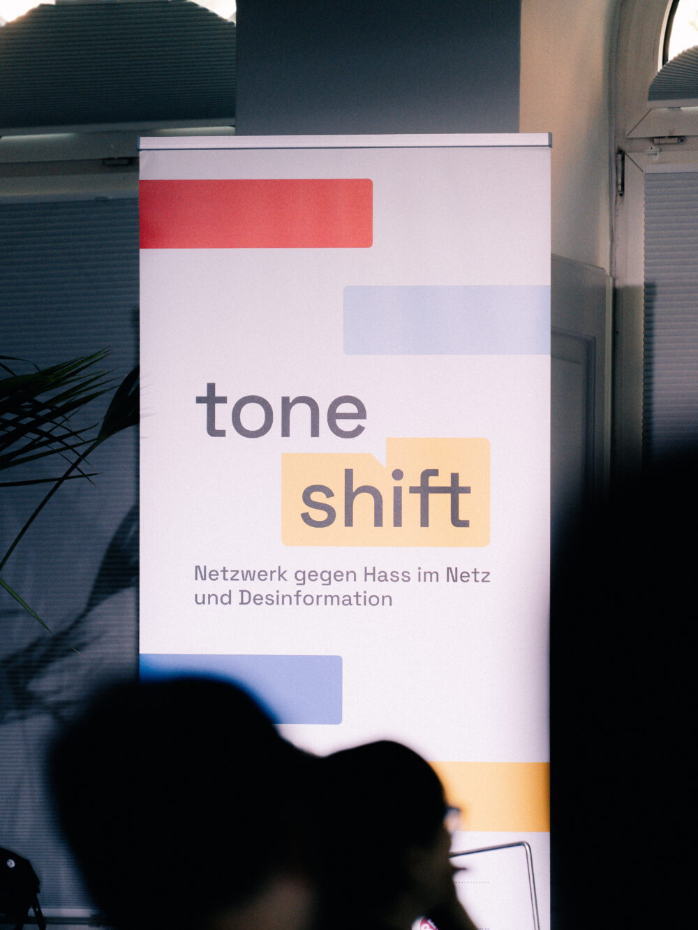 Aufsteller mit dem Schriftzug 'tone shift' und dem Text 'Netzwerk gegen Hass im Netz und Desinformation' vor einer Wand mit Fenster und Pflanze im Hintergrund.