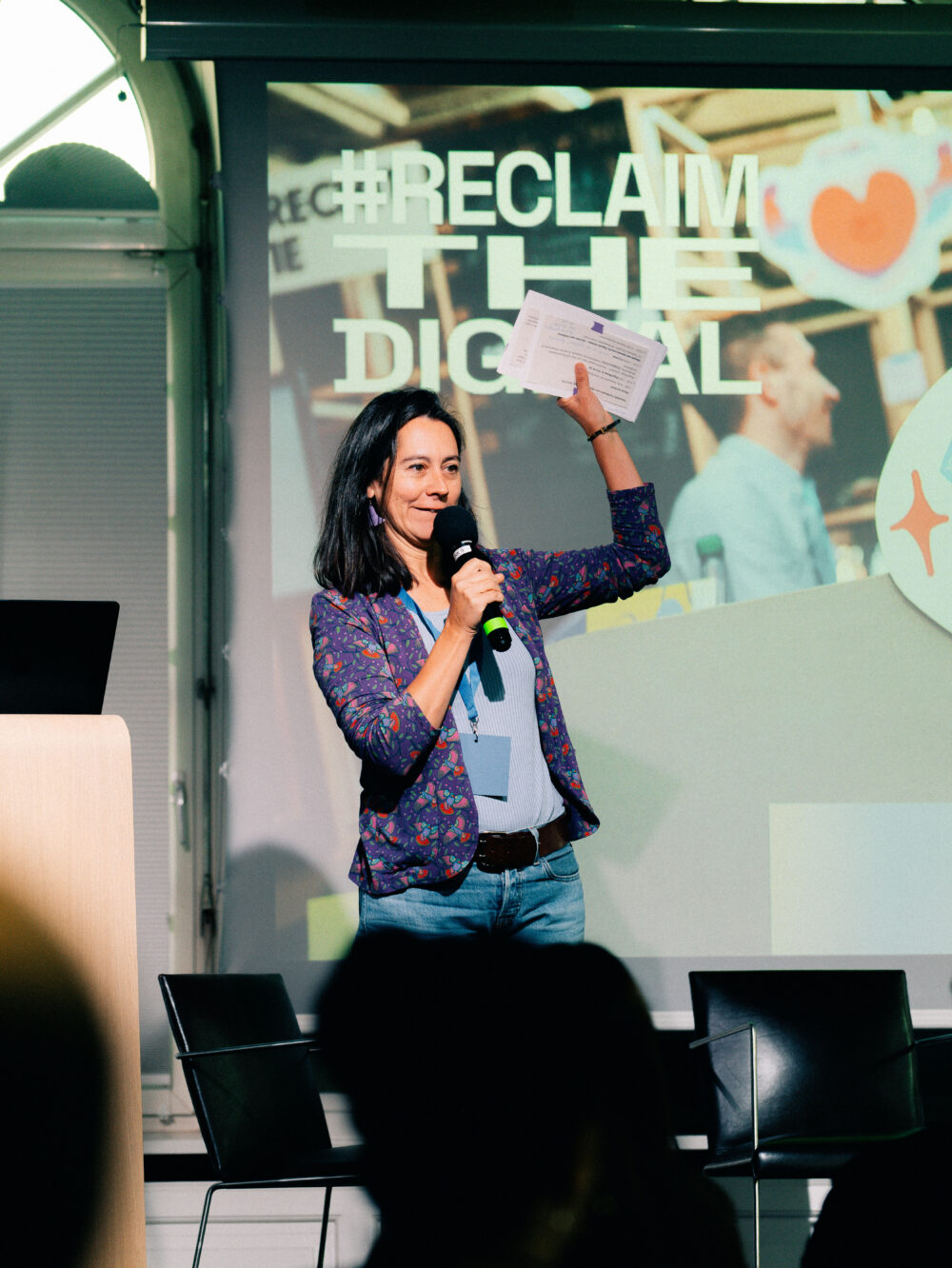Frau mit Mikrofon steht vor Leinwand mit Schriftzug '#RECLAIM THE DIGITAL' und spricht in einem Veranstaltungsraum.