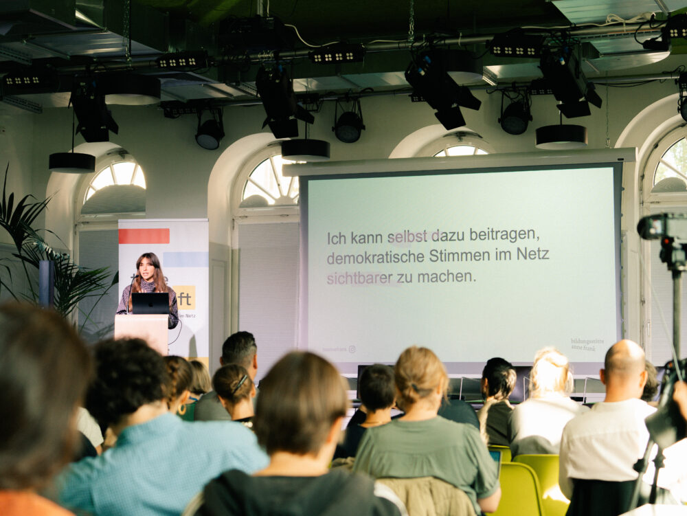 Frau steht am Rednerpult vor Publikum in hellem Raum mit großen Fenstern und präsentiert Text auf Leinwand.