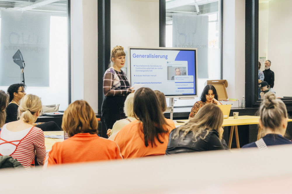 Zwei Personen halten einen Vortrag: Eine steht neben einem Bildschirm mit laufender Präsentation, die andere sitzt an einem Tisch davor; im Raum sitzen mehrere Teilnehmende mit dem Rücken zur Kamera.