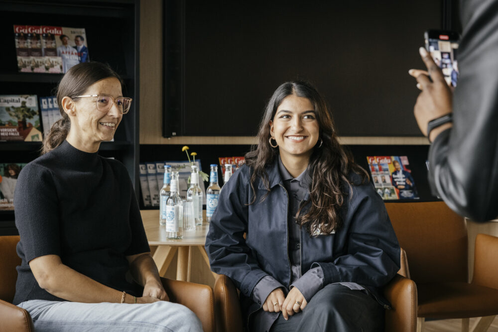 Das Bild zeigt zwei sitzende Personen, die von einer Person mit einem Handy fotografiert werden, dabei lächeln sie.