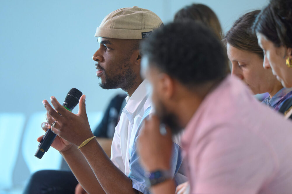 Das Bild zeigt einen Mann von der Seite, der auf einer Veranstaltung in ein Mikrofon spricht. Neben ihm sitzen weitere Personen.