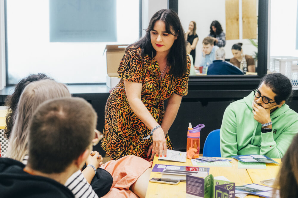 Auf dem Bild sieht man Personen, die an einem Tisch sitzen auf dem verschiedene Materialien liegen. Zwei Personen sitzen mit dem Rücken zum Fotografen. Eine Person mit langen Haaren steht und erklärt etwas.