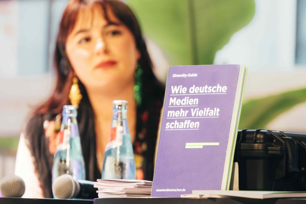 Das Bild zeigt die Publikation "Diversity-Guide – Wie deutsche Medien mehr Vielfalt schaffen" der Neuen deutschen Medienmacher*innen. Die Publikation steht bei einer Veranstaltung auf einem Tisch. Dahinter sitzt eine Person.