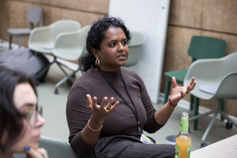 Eine Frau sitzt vor einem Tisch, mit einem Getränk. Sie spricht und gestikuliert mit den Händen. Rechts neben ihr ist eine weitere Person unscharf zu sehen. Die Szene zeigt eine Workshopsituation.