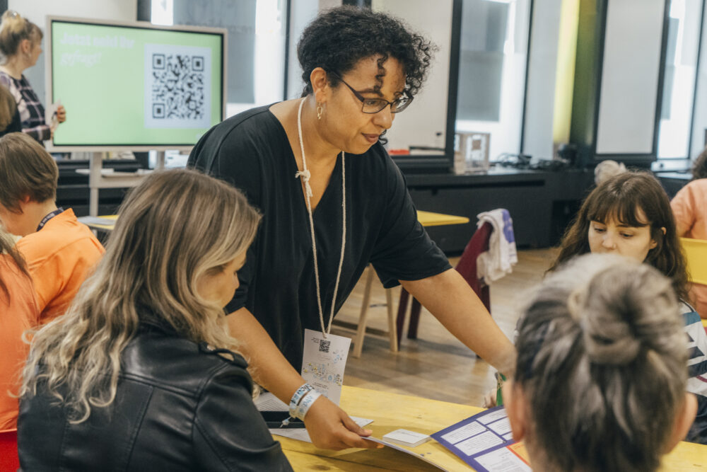 Das Foto zeigt eine Workshopsituation. Eine Person steht vor einem Tisch, um den drei Personen sitzen. Sie hält Arbeitsmaterialien in ihren Händen, die sie den am Tisch sitzenden Personen zeigt. Im Hintergrund steht ein großer Bildschirm mit einem QR-Code.